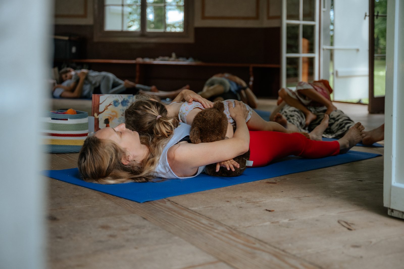 Eine Mutter liegt mit ihrem Kind auf der Yogamatte, das Kind hält einen Stoffbären, beide ruhen.